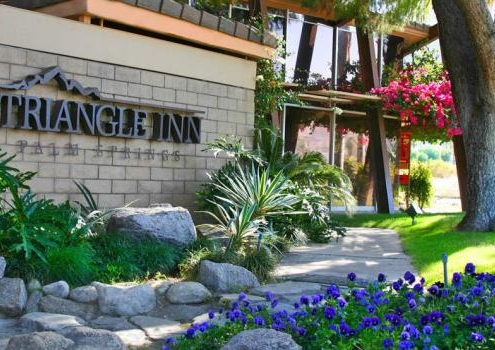 Blue flowers are planted in front of the welcoming Triangle Inn Palm Springs sign at the entrance to this men's clothing-optional resort