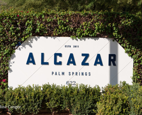 The white sign at the front of Alcazar Palm Springs with blue letters spelling out the hotel's name