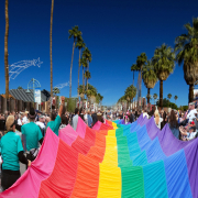 People wearing turquoise shirts carry a large rainbow flag down a palm tree lined street in Palm Springs