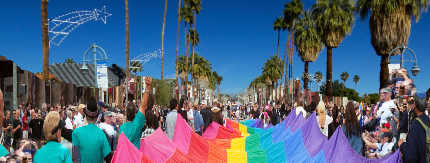 People wearing turquoise shirts carry a large rainbow flag down a palm tree lined street in Palm Springs