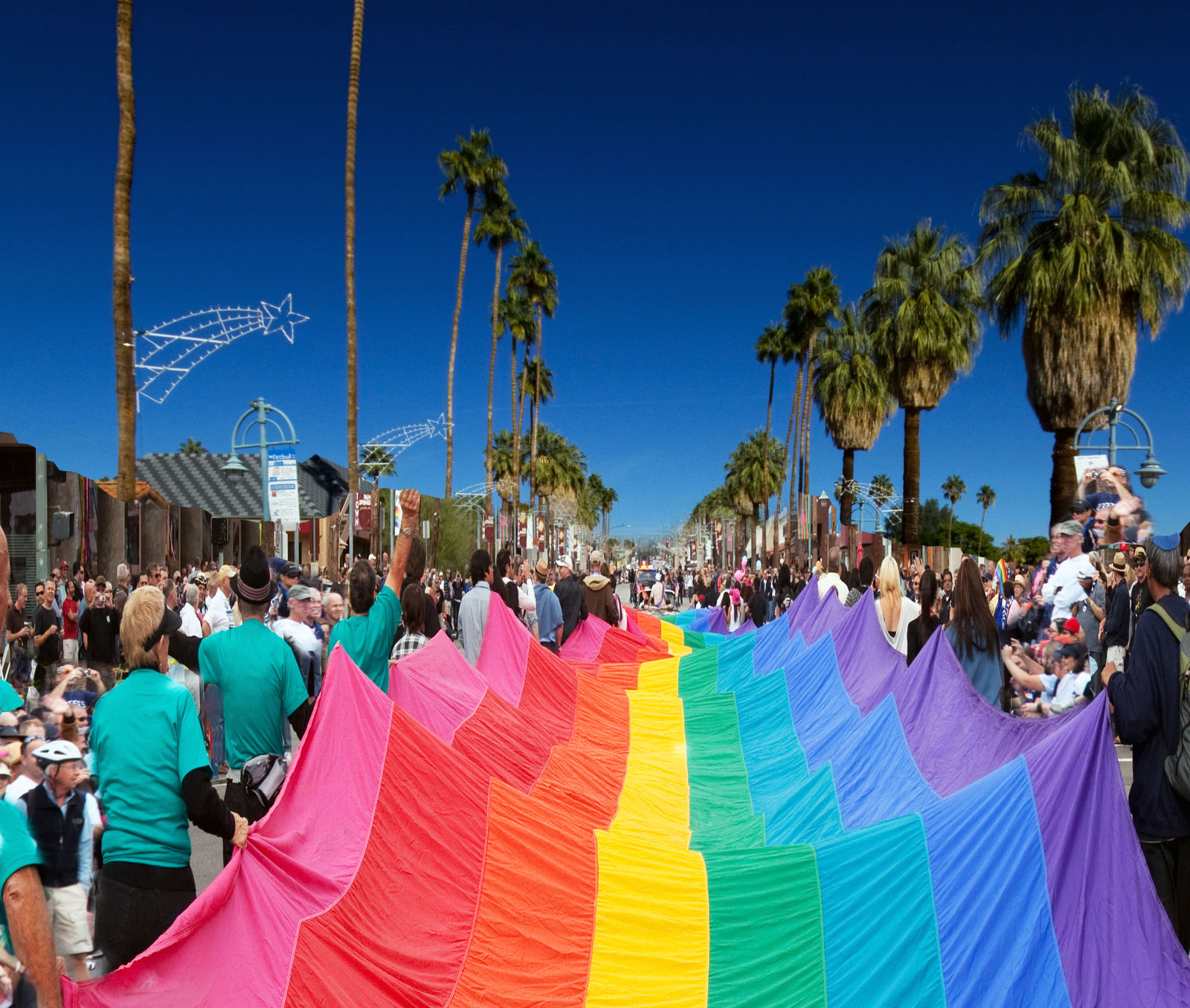 People wearing turquoise shirts carry a large rainbow flag down a palm tree lined street in Palm Springs