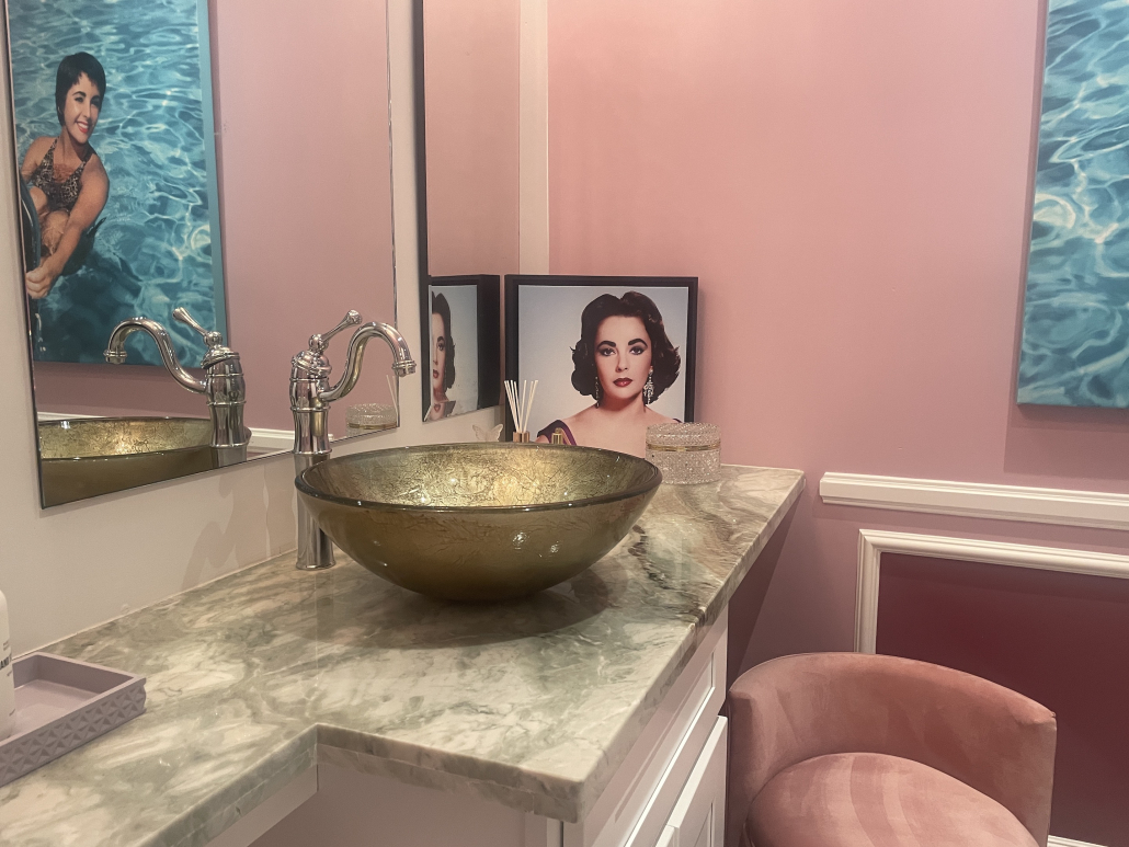 Inside a bathroom with pink painted walls and a gold sink and a photo of Elizabeth Taylor in a black frame