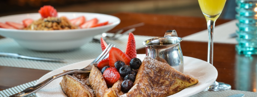 A white plate with French toast and berries on top of it with a mimosa in a glass next to it served at Escena