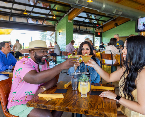 A Black man, a white woman, and a Hispanic woman hold their glasses up to cheers at a table