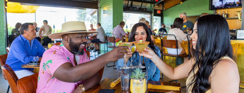 A Black man, a white woman, and a Hispanic woman hold their glasses up to cheers at a table