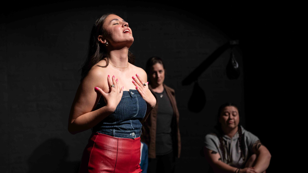 A woman in a black top and red skirt sings while on a dark stage