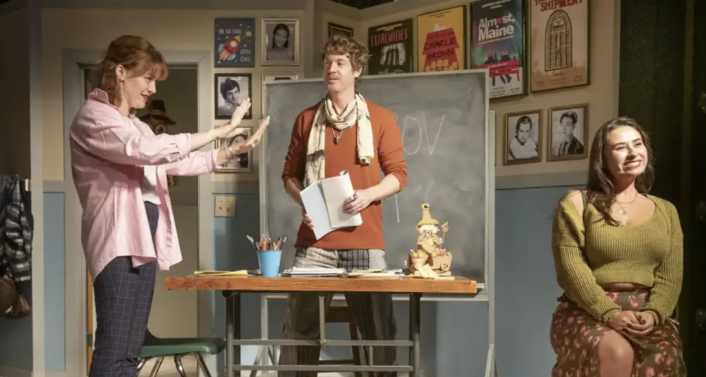 A white woman stands next to a white man holding a book while a smiling white woman sits down in a chair