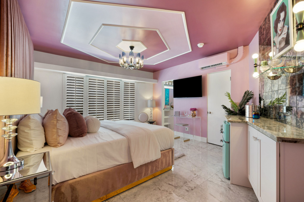 A bed under a crystal light fixture with a lavender wall and white shutters on the windows