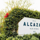 A white sign that says Alcazar Palm Springs in blue letters surrounded by green shrubbery